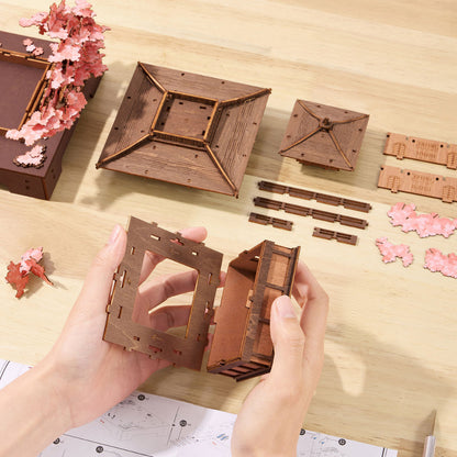 A pair of hands assembling a wooden 3D model of a traditional Japanese pagoda on a light wood tabletop, with other pre-cut wooden pieces and decorative pink cherry blossom ornaments arranged around it.