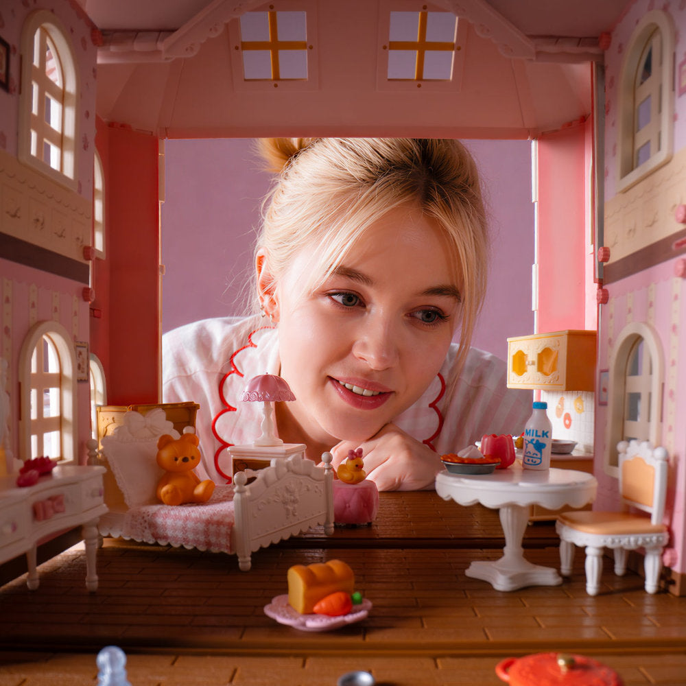 A young blonde girl with her hair in a bun smiles while looking down at a miniature dollhouse interior. The dollhouse features pink walls, tiny beds, a small dining table with chairs, and toy furniture, creating a cozy and playful scene.