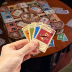 A hand holds a fan of colorful game cards in front of a wooden table with a board game setup spread out in the background.