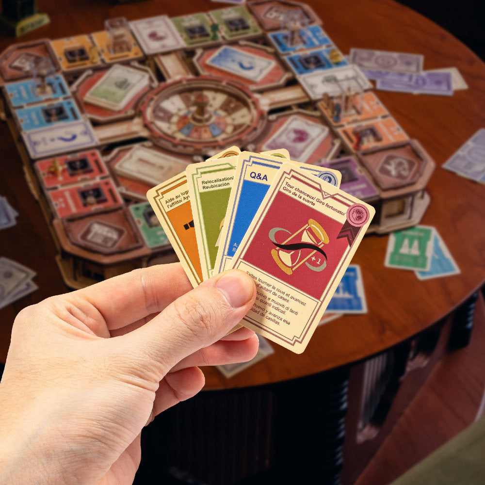 A hand holds a fan of colorful game cards in front of a wooden table with a board game setup spread out in the background.