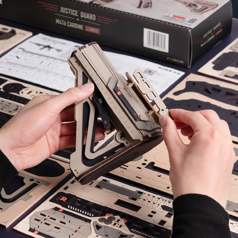 A person's hands are assembling a wooden 3D model of a firearm, holding a layered wooden piece over a sheet of corresponding laser-cut wood pieces. The product box labeled 'JUSTICE GUARD MA14 CARRINE' is visible in the background along with other assembly sheets.