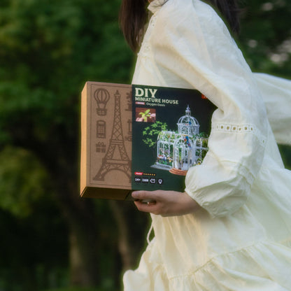 A person in a white dress holds a DIY miniature house kit box outdoors, with a blurred green background.