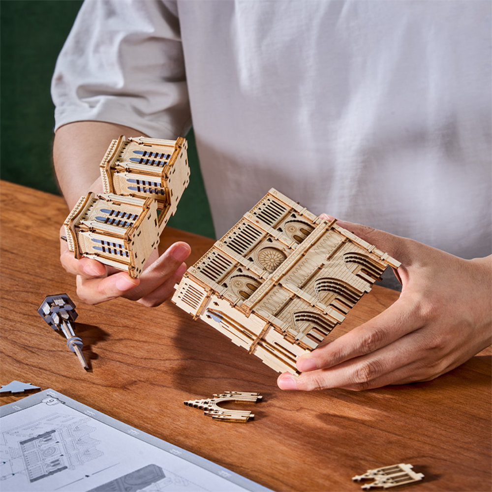 Person assembling a wooden mechanical model on a wooden table with a white shirt.