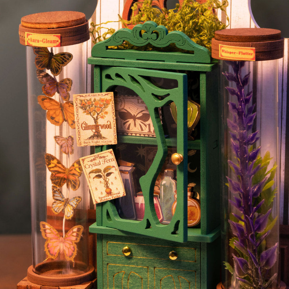 A miniature green display cabinet with ornate carvings and brass-tone knobs, styled with small potion bottles, vintage-style labels, and two large glass specimen jars containing preserved butterflies and purple flowers.