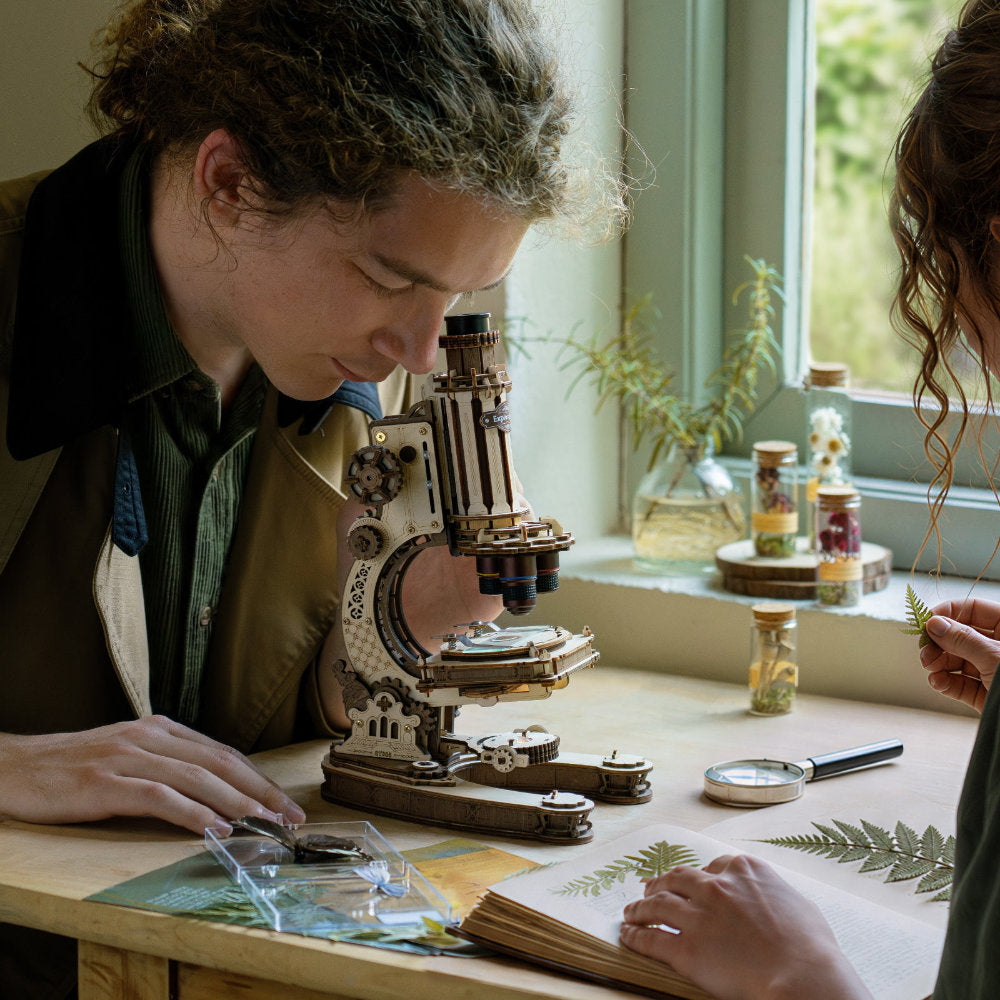 Two people examining plants under a microscope in a room with a window and plants.
