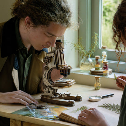 Two people examining plants under a microscope in a room with a window and plants.