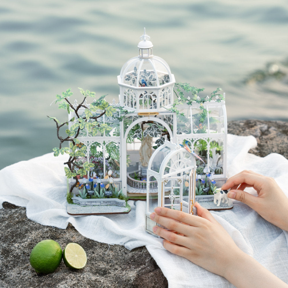 A miniature white glass-domed garden pavilion model with tiny trees, furniture, and small figurines, displayed on a white cloth over rocks with a blurred body of water in the background. Two hands are assembling or adjusting the model, and two green limes sit nearby.