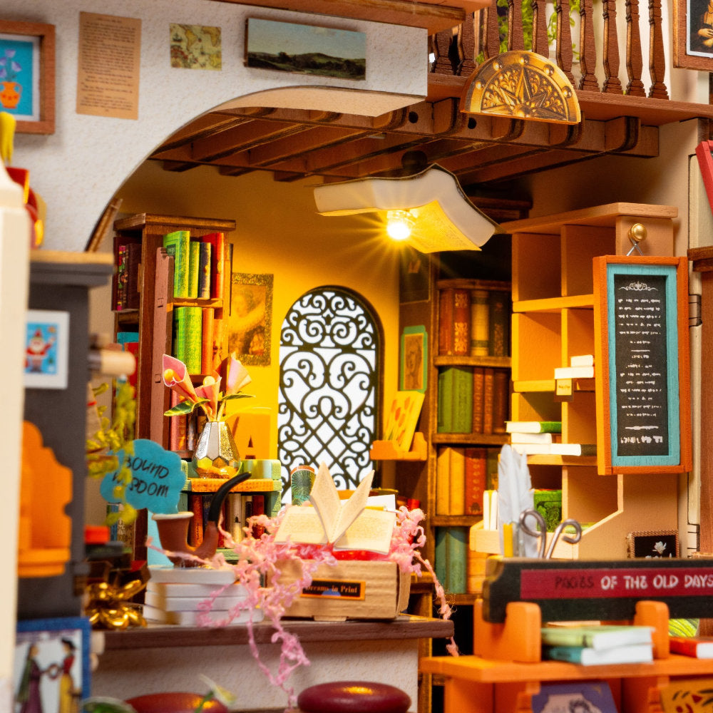 A miniature bookshop dollhouse interior with tiny bookshelves, a chalkboard, hanging lanterns, and warm yellow lighting, photographed as a product scene rather than a real environment.