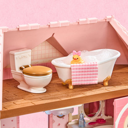 A pink dollhouse-style bathroom set with a white clawfoot bathtub filled with bubbles and a yellow rubber duck, alongside a white toilet with a gold lid on a wooden floor against a pink wall.