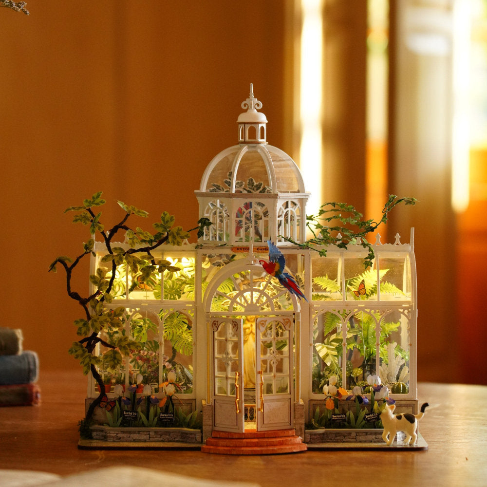A miniature greenhouse-style decorative model with glowing warm lights, small plants, flowers, a bird perched on the roof, and a small dog figurine, displayed on a wooden surface against a softly blurred warm indoor background.
