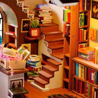 A miniature dollhouse-style interior scene with a wooden staircase, bookshelves, and many colorful books. The room is highly detailed and styled like a cozy library, with small furniture pieces, framed pictures, and decorative items.