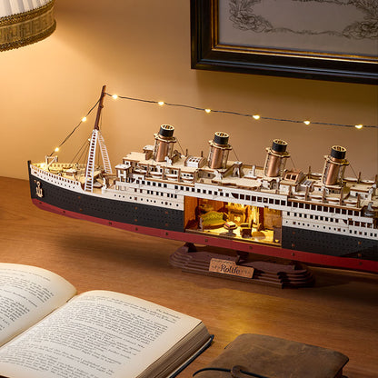 Model of a ship on a wooden surface with an open book and framed picture in the background.