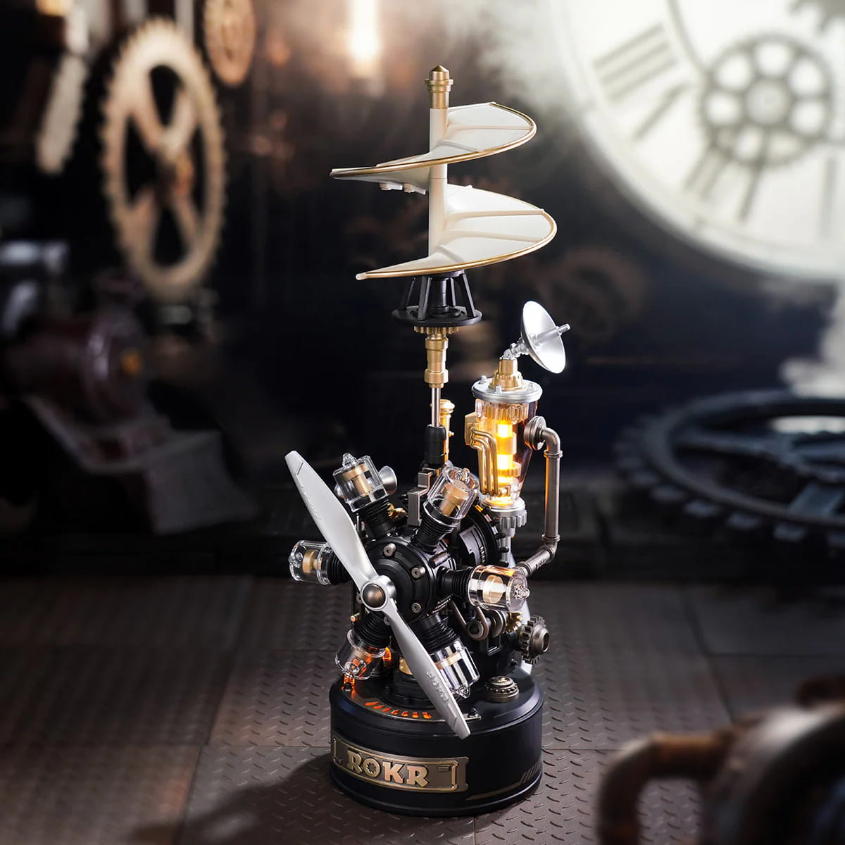 Steampunk-style decorative model on a round black base with a spiral propeller, metal gears, a small glowing amber chamber, and a white dish antenna, photographed against a dark industrial background with blurred clock and gear elements.