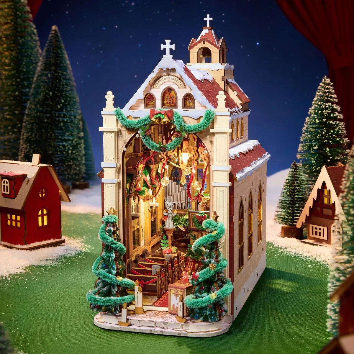 Wooden-Town Caroling Cathedral 3D wooden book nook kit – cathedral with lit interior on a snowy green base beside small houses and trees